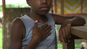 Young girl from Vanuatu leaning on a railing looking at the camera with her hand resting on her chest over her heart