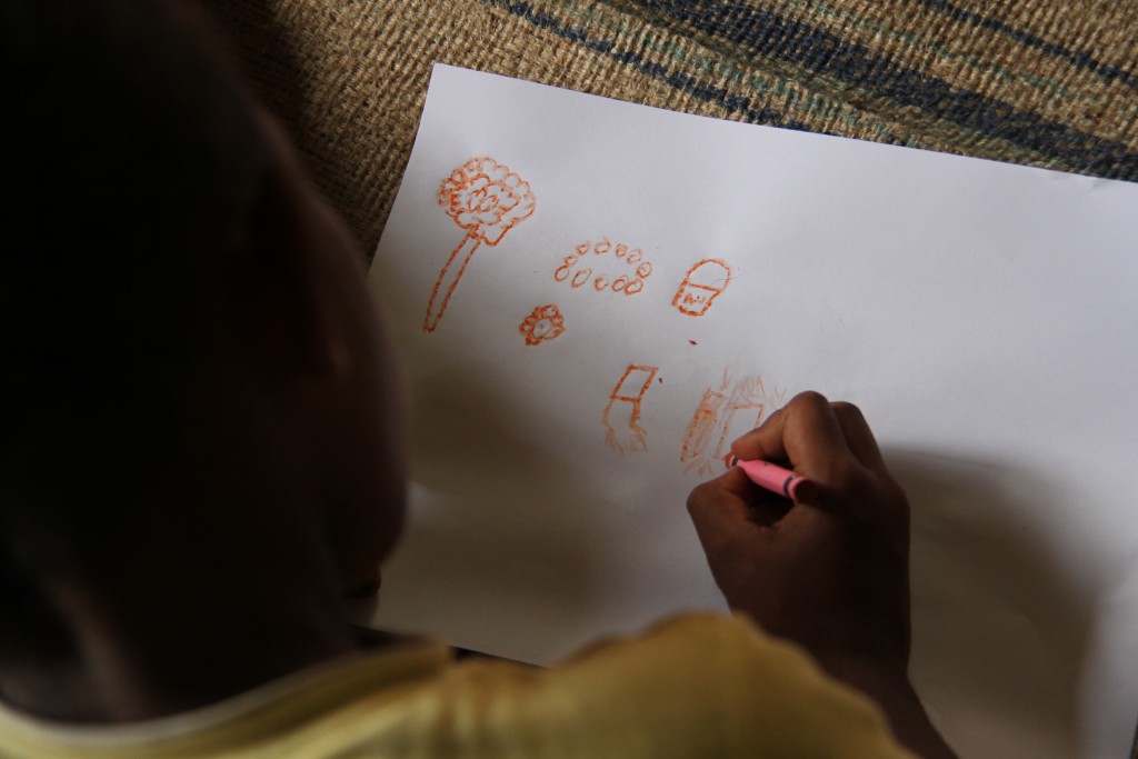 A photo taken looking over the shoulder of a child lying on a mat drawing a picture